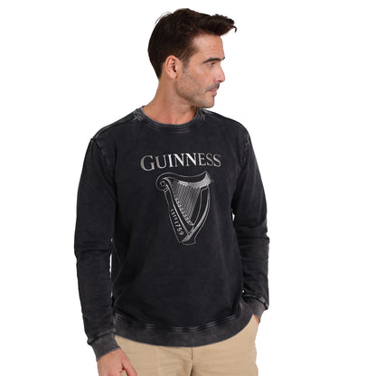 A man stands facing right, one hand in his pocket, wearing the Guinness Embossed Print Trademark Sweater from Guinness Webstore US with a faded black finish and harp logo for a vintage look.