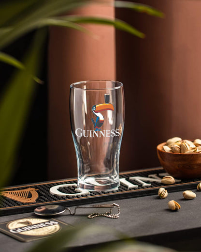 A Guinness Toucan Pint Glass by Guinness sits on a bar mat with pistachios and a bowl nearby, framed by plants and curtains in the background.