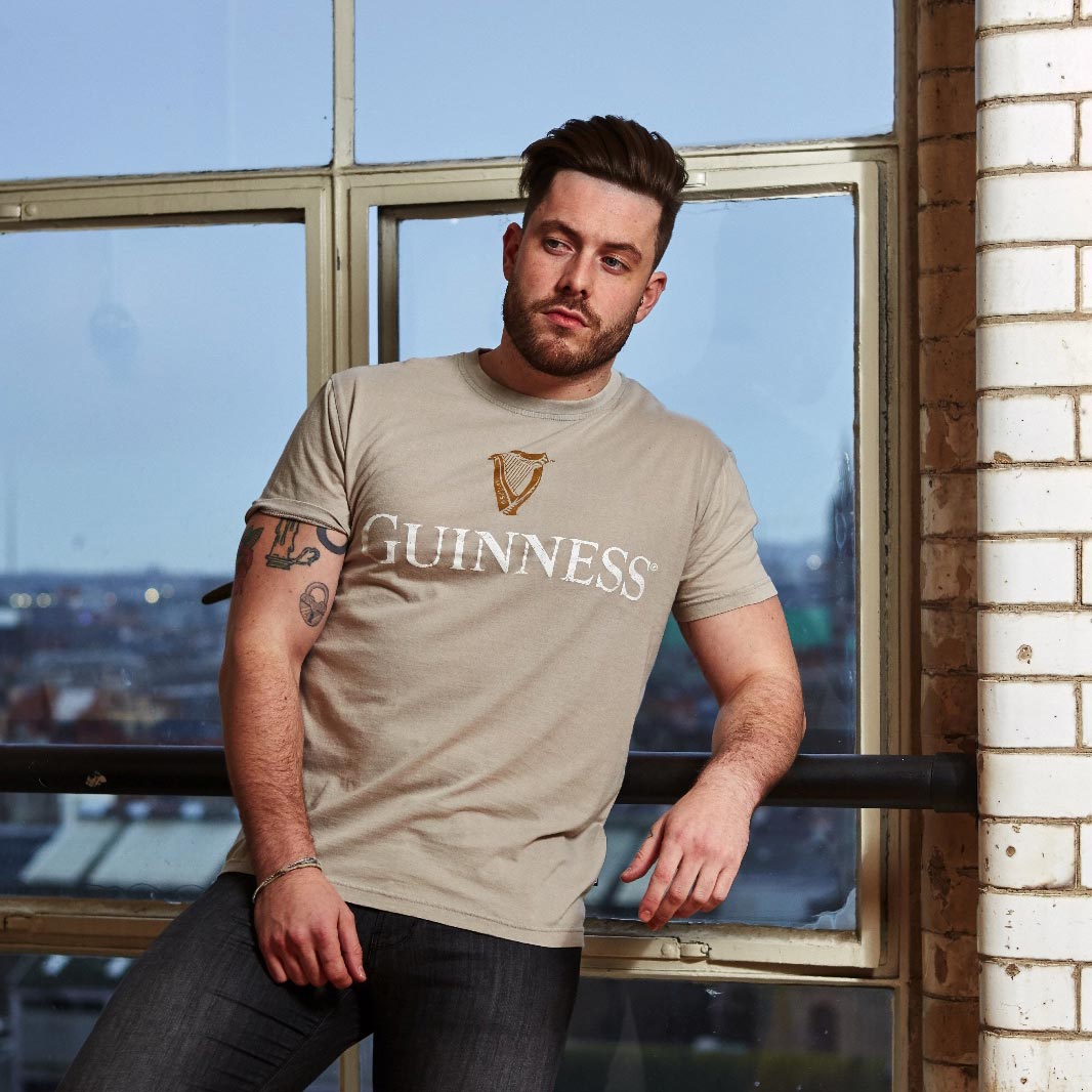 A man wearing a cotton Guinness Trademark Label T-Shirt Beige leaning against a window.