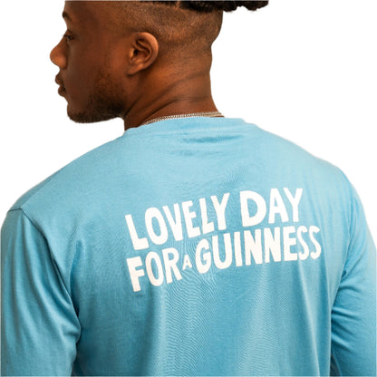 Someone is wearing the Guinness Seb Curi Lovely Day Longsleeve Tee - Blue from Guinness Webstore US, featuring "LOVELY DAY FOR A GUINNESS" printed in white on the back.