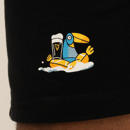 The Guinness Seb Curi Beach Short - Black from Guinness Webstore US features a Sebastian Curi-inspired blue and yellow toucan sitting in foam beside a pint of dark beer, illustrated and printed on black fabric.