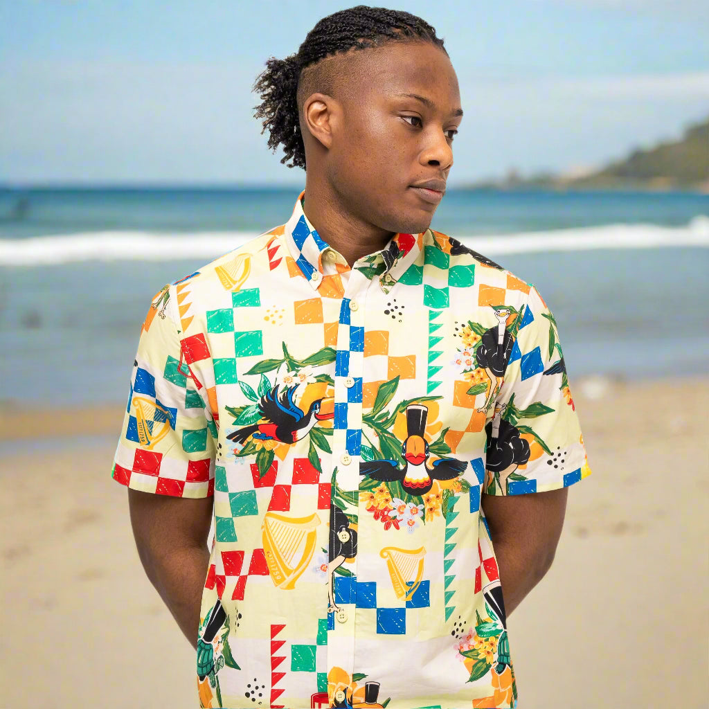A person stands on a beach wearing the Guinness Toucan Beach Shirt from Guinness Webstore US, its colorful woven cotton design visible as they look to the side with the ocean and shoreline in the background.