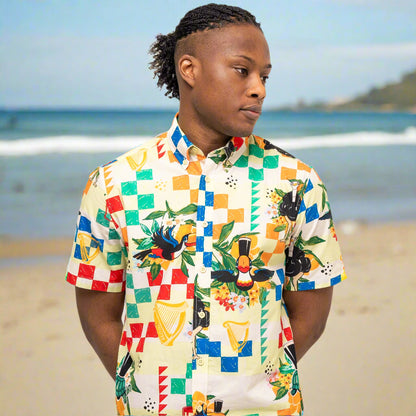 A person stands on a beach wearing the Guinness Toucan Beach Shirt from Guinness Webstore US, its colorful woven cotton design visible as they look to the side with the ocean and shoreline in the background.