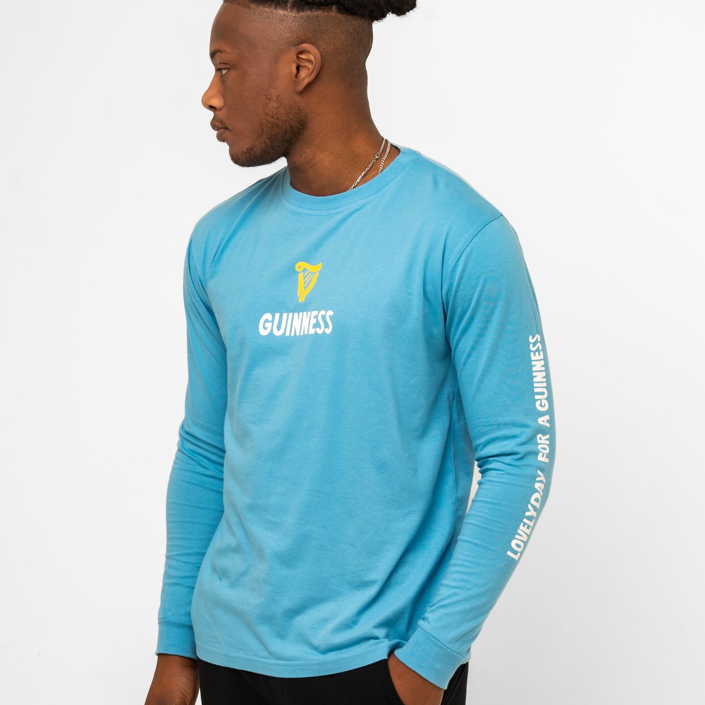 A man models the Guinness Seb Curi Lovely Day Longsleeve Tee in blue from Guinness Webstore US, featuring "GUINNESS" and its logo on the chest and "LOVELY DAY FOR A GUINNESS" on the sleeve, against a white background.