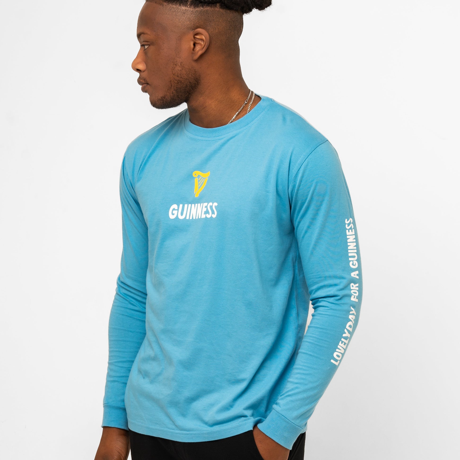 A man models the Guinness Seb Curi Lovely Day Longsleeve Tee in blue from Guinness Webstore US, featuring "GUINNESS" and its logo on the chest and "LOVELY DAY FOR A GUINNESS" on the sleeve, against a white background.