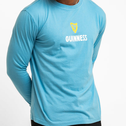 A person wears the Guinness Seb Curi Lovely Day Longsleeve Tee - Blue from Guinness Webstore US, a pop-art tee with a yellow harp and "GUINNESS" graphic on the front, set against a plain white background.