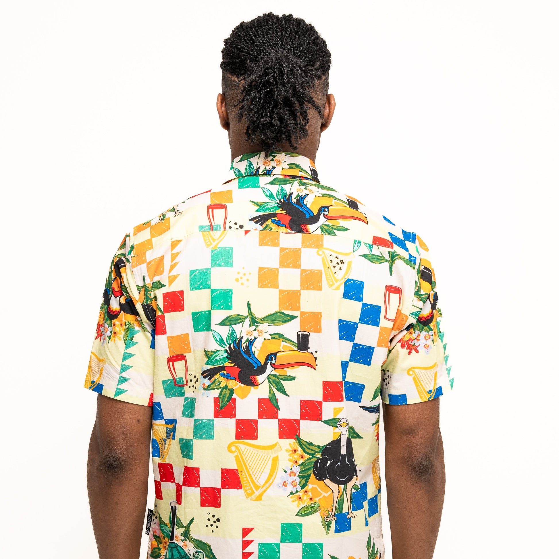 A person with braided hair is seen from behind wearing the Guinness Toucan Beach Shirt by Guinness Webstore US, a vibrant short-sleeve shirt with breathable woven cotton and lively illustrations including checkerboard designs.