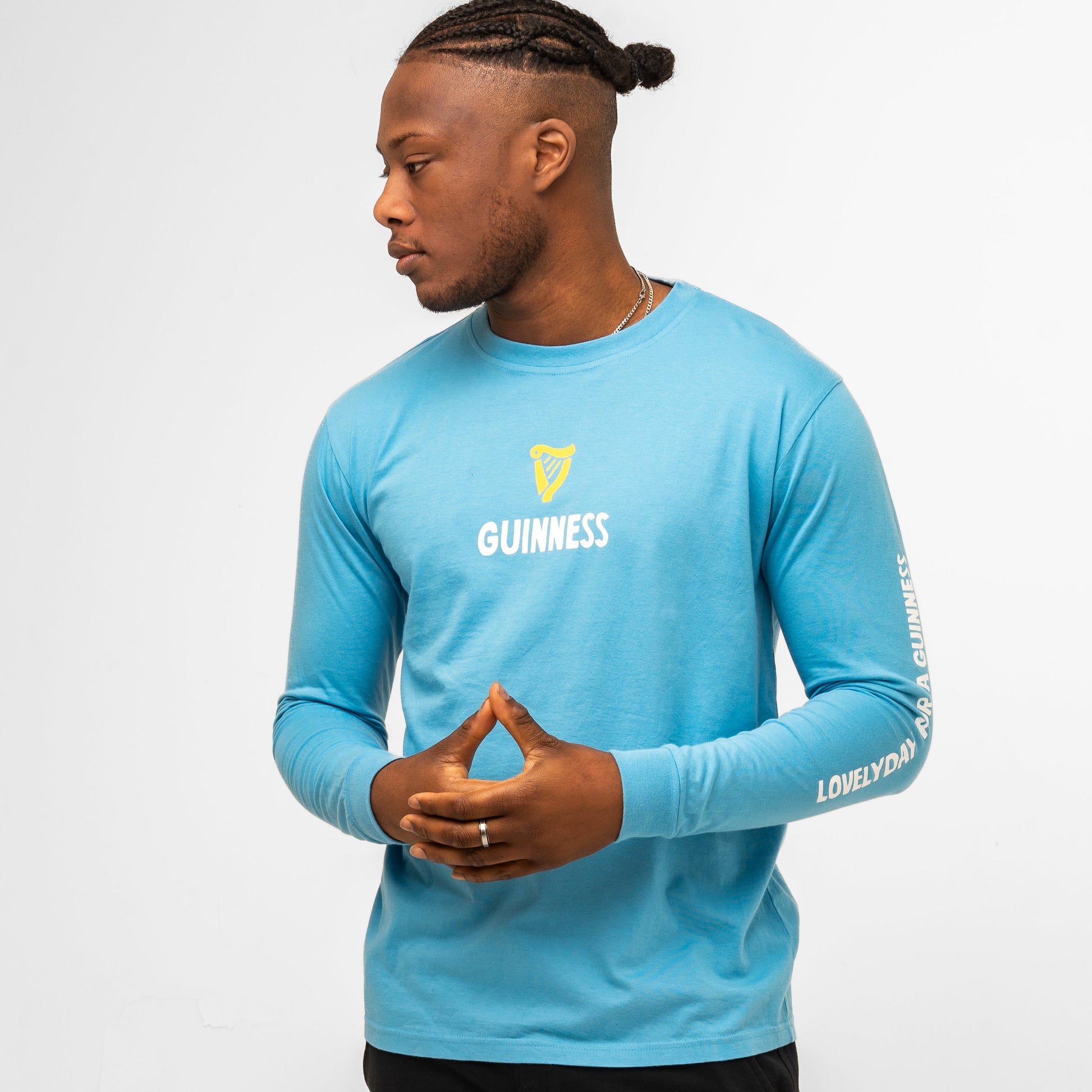 A man in a light blue Guinness Seb Curi Lovely Day Longsleeve Tee by Guinness Webstore US, featuring the harp logo and pop-art graphics from the Sebastian Curi collaboration, stands against a white background, looking left with hands clasped.