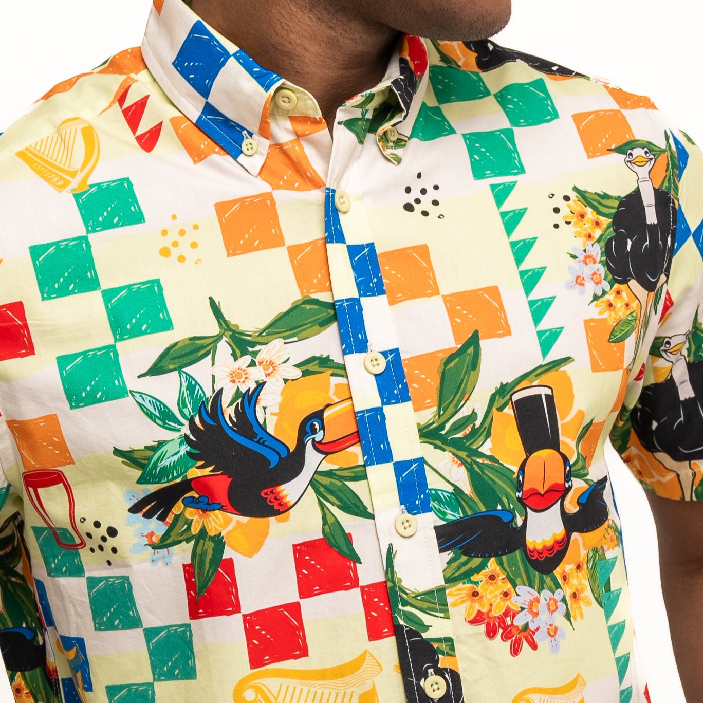 A person wears the Guinness Toucan Beach Shirt from Guinness Webstore US—a Hawaiian-style short-sleeve shirt with a colorful checkered pattern, tropical birds, flowers, and musical instruments.