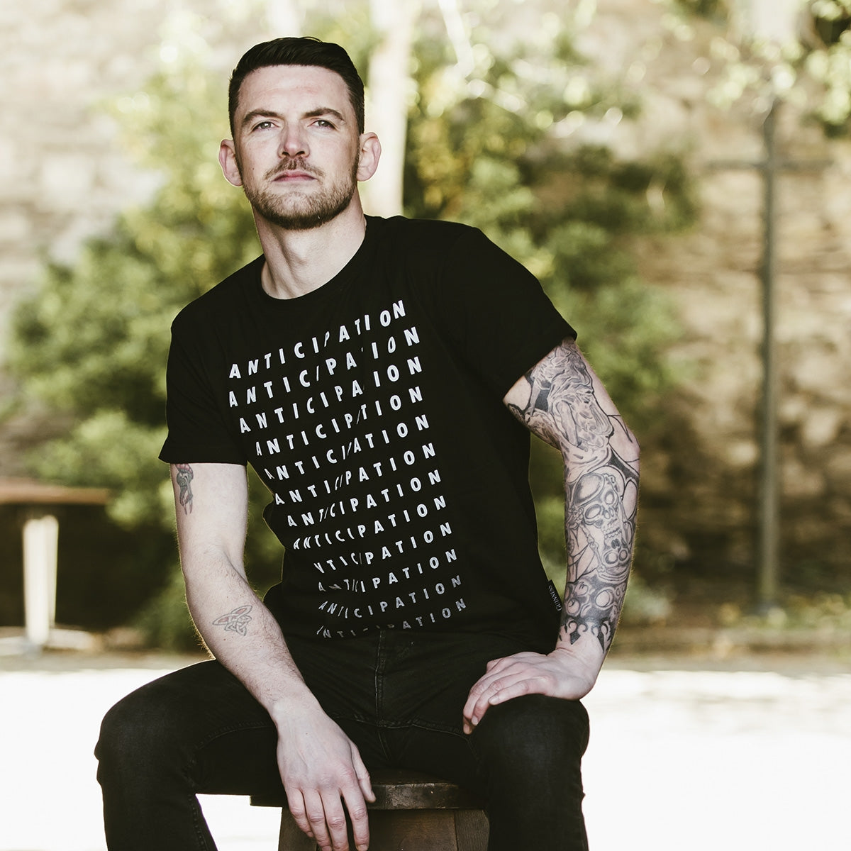 A man sitting on a stool with tattoos, displaying both comfort and style in his Guinness Anticipation Pint tee from Guinness.