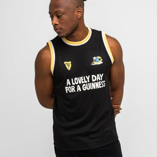 A man wears the Guinness Seb Curi Basketball Jersey from Guinness Webstore US—a black limited-edition jersey with yellow and white trim reading "A LOVELY DAY FOR A GUINNESS"—while standing against a plain background.