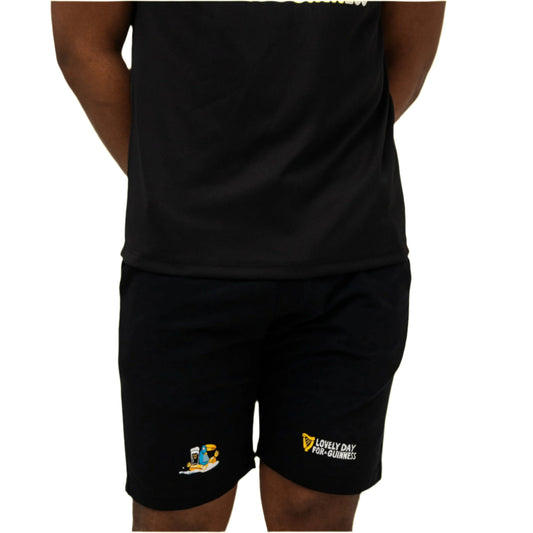 A person stands with hands behind their back, wearing a black t-shirt and the Guinness Seb Curi Beach Short - Black from Guinness Webstore US, featuring small yellow and blue graphics and text near the left knee.