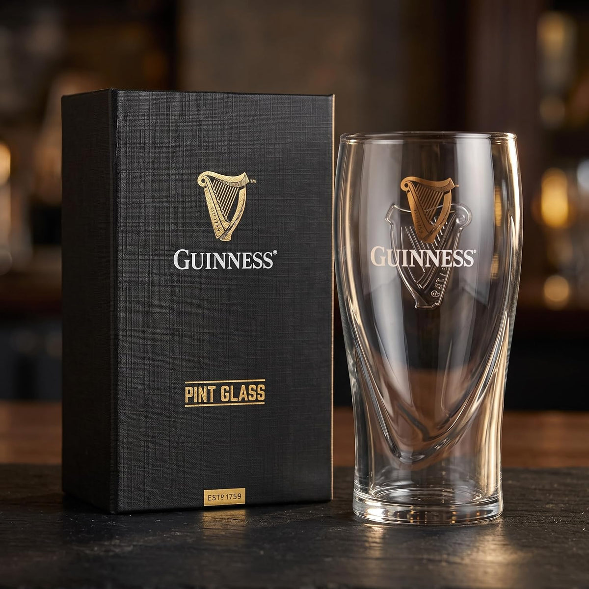 A Guinness® Pint Glass with Gift Box featuring a harp logo standing next to its black presentation box.
