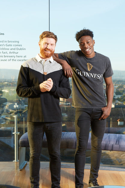 Two men wearing Guinness Heritage Charcoal Grey & Black Long-Sleeve Rugby jerseys standing next to each other in front of a building.