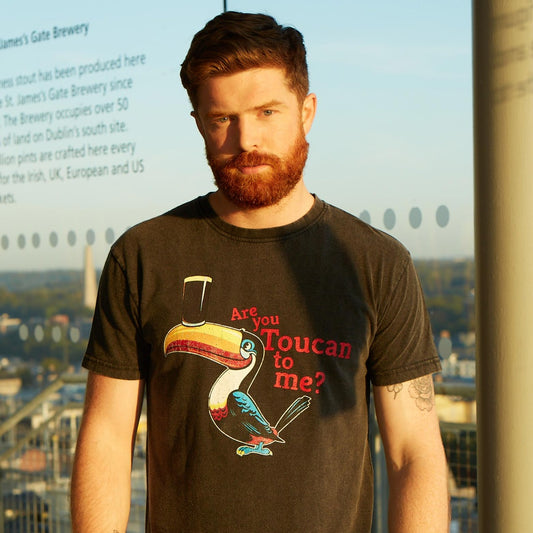 A man with a red beard stands outdoors wearing the Guinness Are You Toucan To Me Tee in grey, featuring a colorful toucan and playful text. The cityscape and glass panels provide a vibrant backdrop to this Guinness shirt.