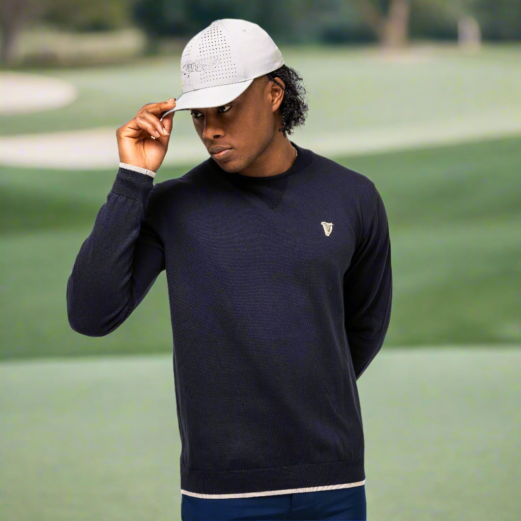 A person in a gray cap, Guinness Performance Cotton Sweater - Navy with harp logo from Guinness Webstore US, and navy shorts stands against a white background, holding the brim of the cap with one hand.