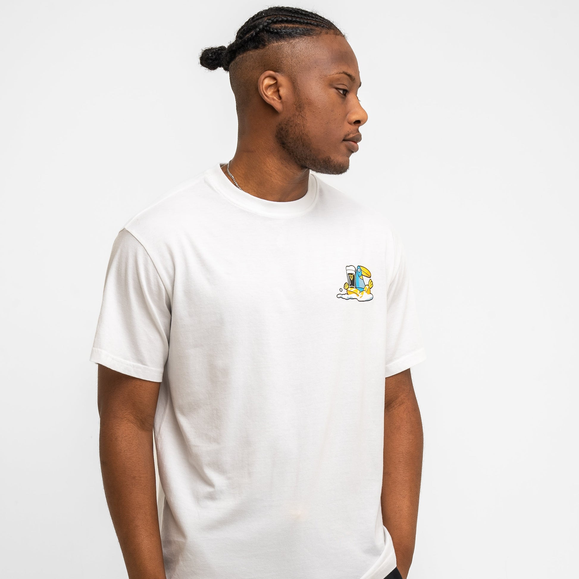 A man with braided hair wears the Guinness Seb Curi Oversize Tee in white from Guinness Webstore US, featuring a small colorful chest graphic, as he stands against a plain white background looking to his left.