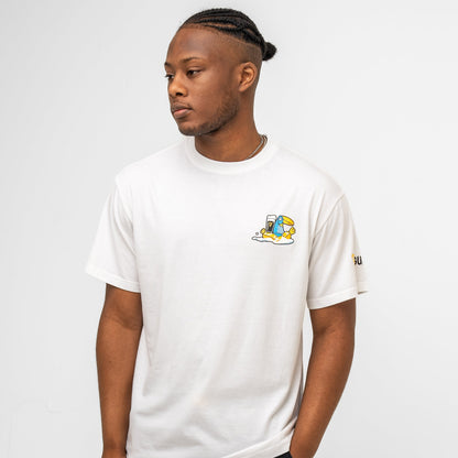 A man with braided hair wears the Guinness Seb Curi Oversize Tee - White from Guinness Webstore US, featuring a small yellow and blue cartoon graphic by Sebastian Curi on the left chest, standing against a plain white background.