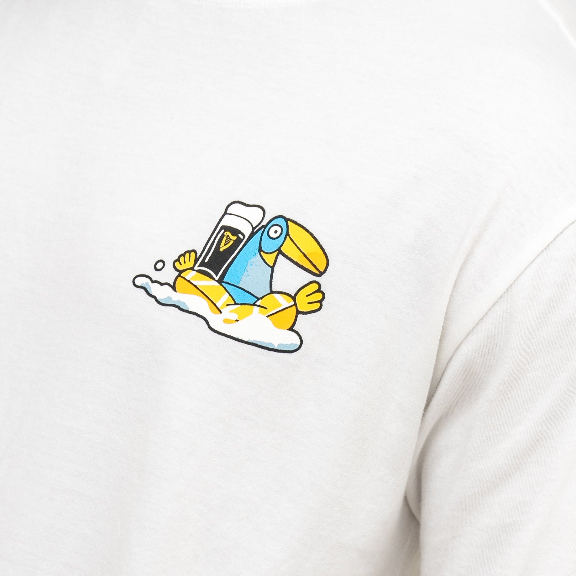The Guinness Seb Curi Oversize Tee - White from Guinness Webstore US showcases a playful blue and yellow toucan relaxing on foam, holding a pint of stout—a vibrant Sebastian Curi collaboration.