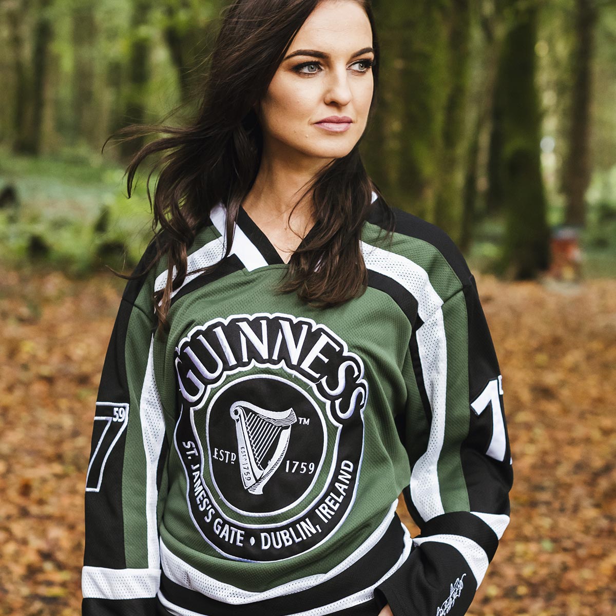 A woman wearing a Guinness Green & White Hockey Jersey poses in the woods.