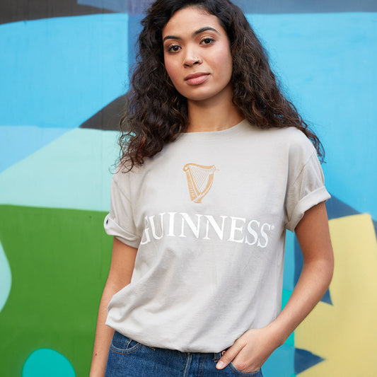 A woman with curly hair is wearing a Beige Guinness Harp Premium Tee by Guinness, standing against a colorful, abstract mural background.