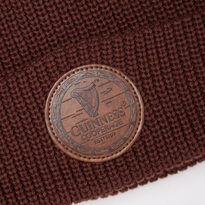 Close-up of the Guinness Cooperage Brown Knitted Beanie with a round leather patch embossed with the cooperage logo and “Guinness Cooperage ESTD 1759” harp design.