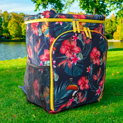 A vibrant, floral-patterned Guinness Toucan Hawaiian Cooler Bag rests on the grass by a lake, basking under the sunny sky, seamlessly blending into nature like outdoor gear from Guinness Webstore US.