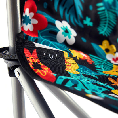 Close-up of the Guinness x Fatti Burke Hawaiian Camping Chair by Guinness, featuring a colorful floral pattern, sturdy metal frame, and a small tag with a smiling face.
