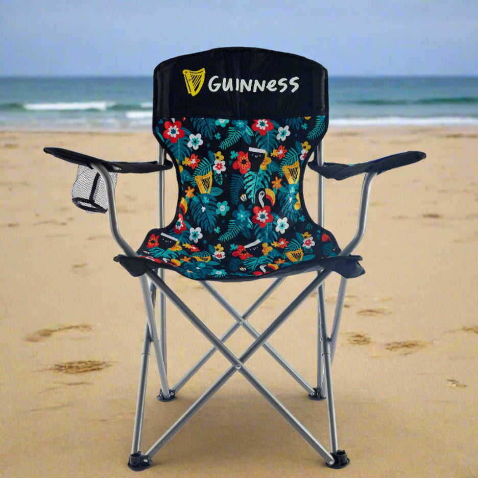 The Guinness x Fatti Burke Hawaiian Camping Chair by Guinness features a floral and logo pattern, armrests, and a mesh cup holder on the left arm, all set against a plain white background.