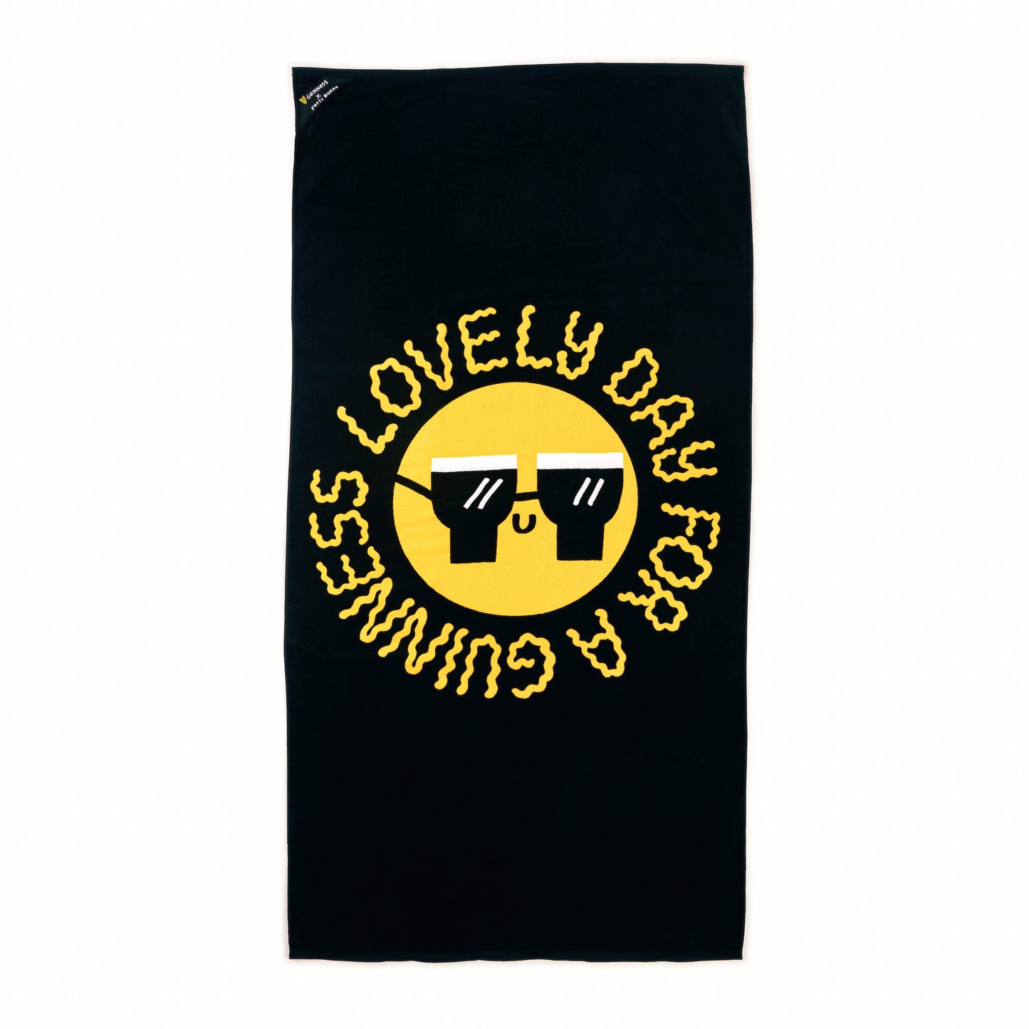A black beach towel featuring a yellow smiley face with sunglasses and the phrase "LOVELY DAY FOR A GUINNESS" in bold yellow letters—perfect for pairing with your Guinness x Fatti Burke Lovely Day Bundle from Guinness Webstore US or cooler bag for a fun day out.