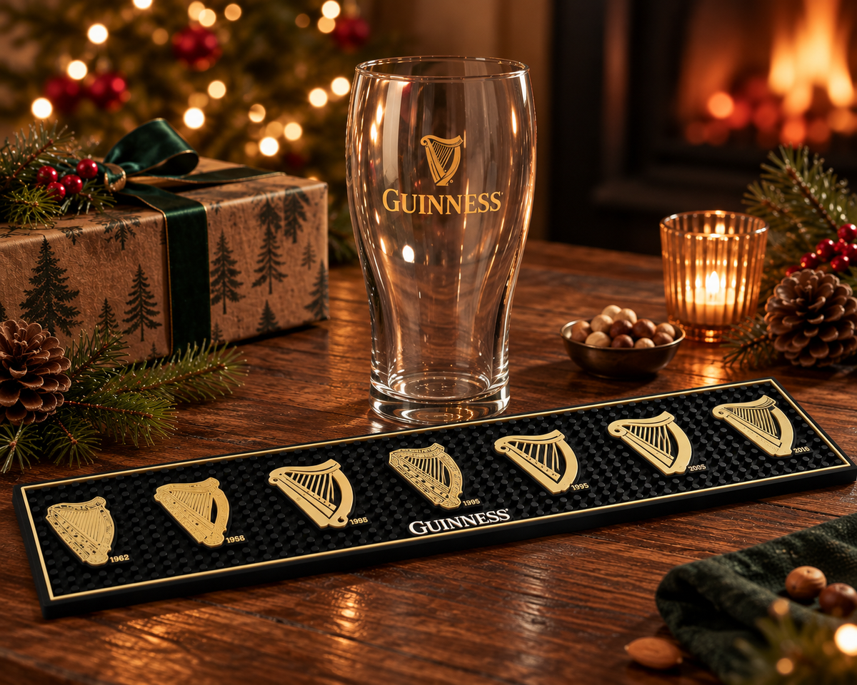 A Guinness pint glass sits behind a black Guinness bar mat featuring six gold-colored harp emblems, part of a Guinness From the Archives Gift Set.
