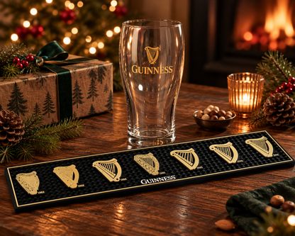 A Guinness pint glass sits behind a black Guinness bar mat featuring six gold-colored harp emblems, part of a Guinness From the Archives Gift Set.