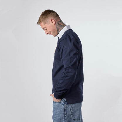 A man with short blond hair and neck tattoos stands in profile against a plain light background, wearing blue jeans and a Guinness Navy Traditional Rugby Jersey from Guinness Webstore US.