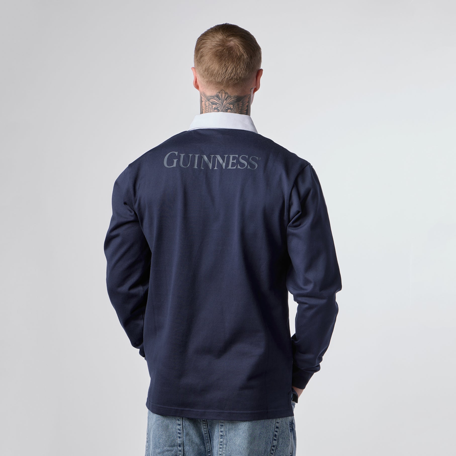 A man with a neck tattoo faces away from the camera, wearing the Guinness Navy Traditional Rugby Jersey from Guinness Webstore US and light blue jeans, set against a plain light background.