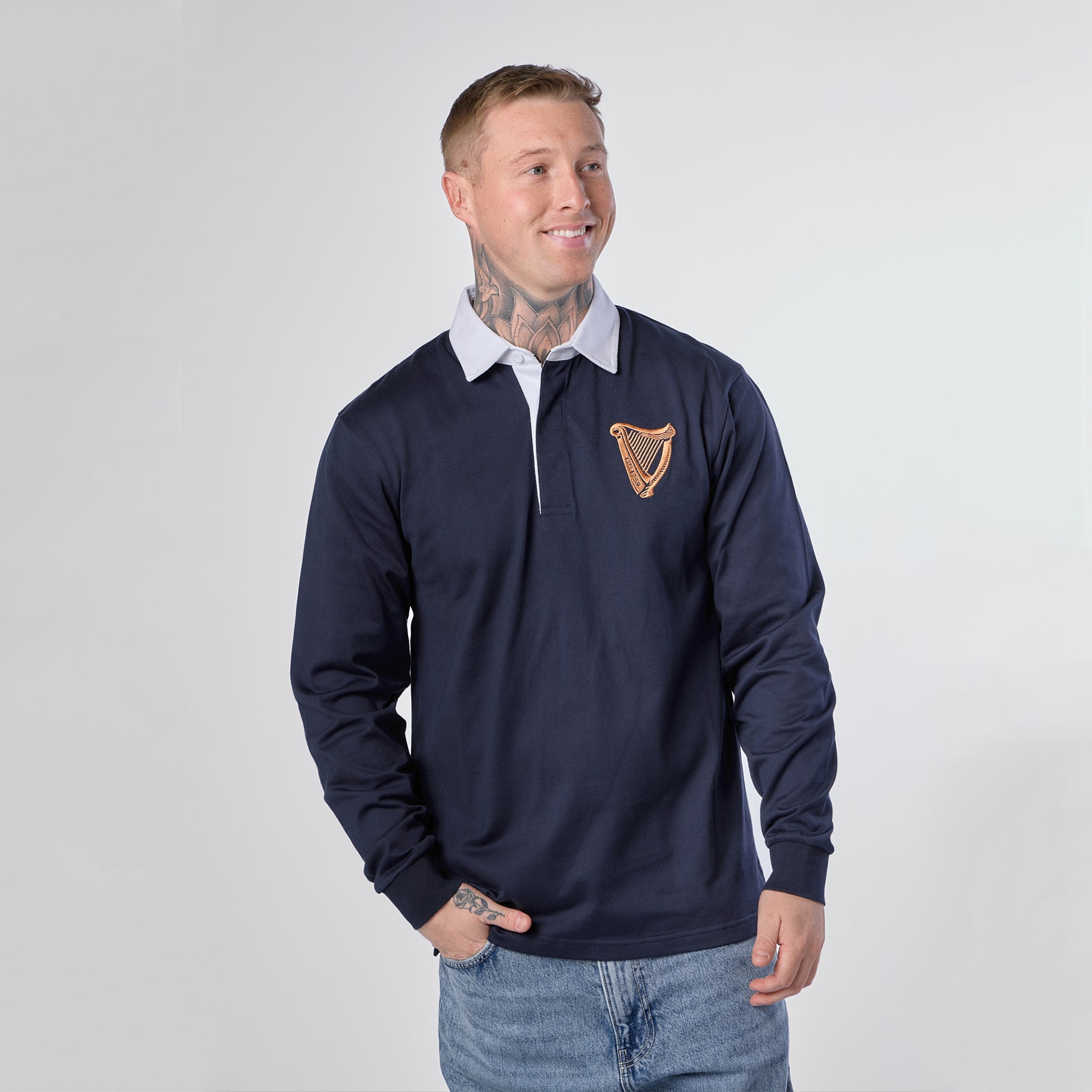 A man wearing the Guinness Navy Traditional Rugby Jersey by Guinness Webstore US—with its signature white collar and gold harp emblem on the chest—stands against a plain light background.