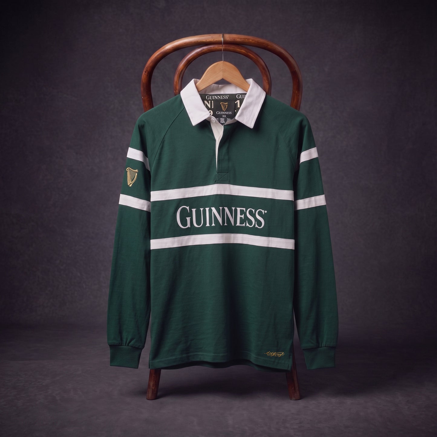 A Guinness Green Traditional Rugby Jersey, featuring white stripes and the iconic “GUINNESS” logo, hangs on a wooden hanger draped over a chair against a dark background.
