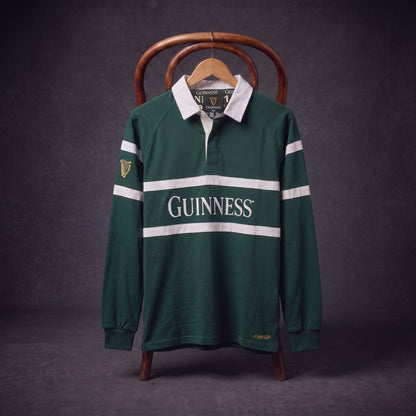 A Guinness Green Traditional Rugby Jersey, featuring white stripes and the iconic “GUINNESS” logo, hangs on a wooden hanger draped over a chair against a dark background.