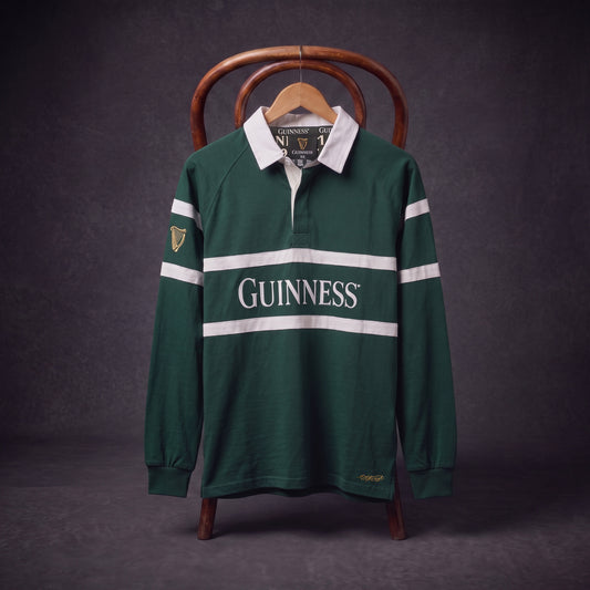 A Guinness Green Traditional Rugby Jersey, featuring white stripes and the iconic “GUINNESS” logo, hangs on a wooden hanger draped over a chair against a dark background.