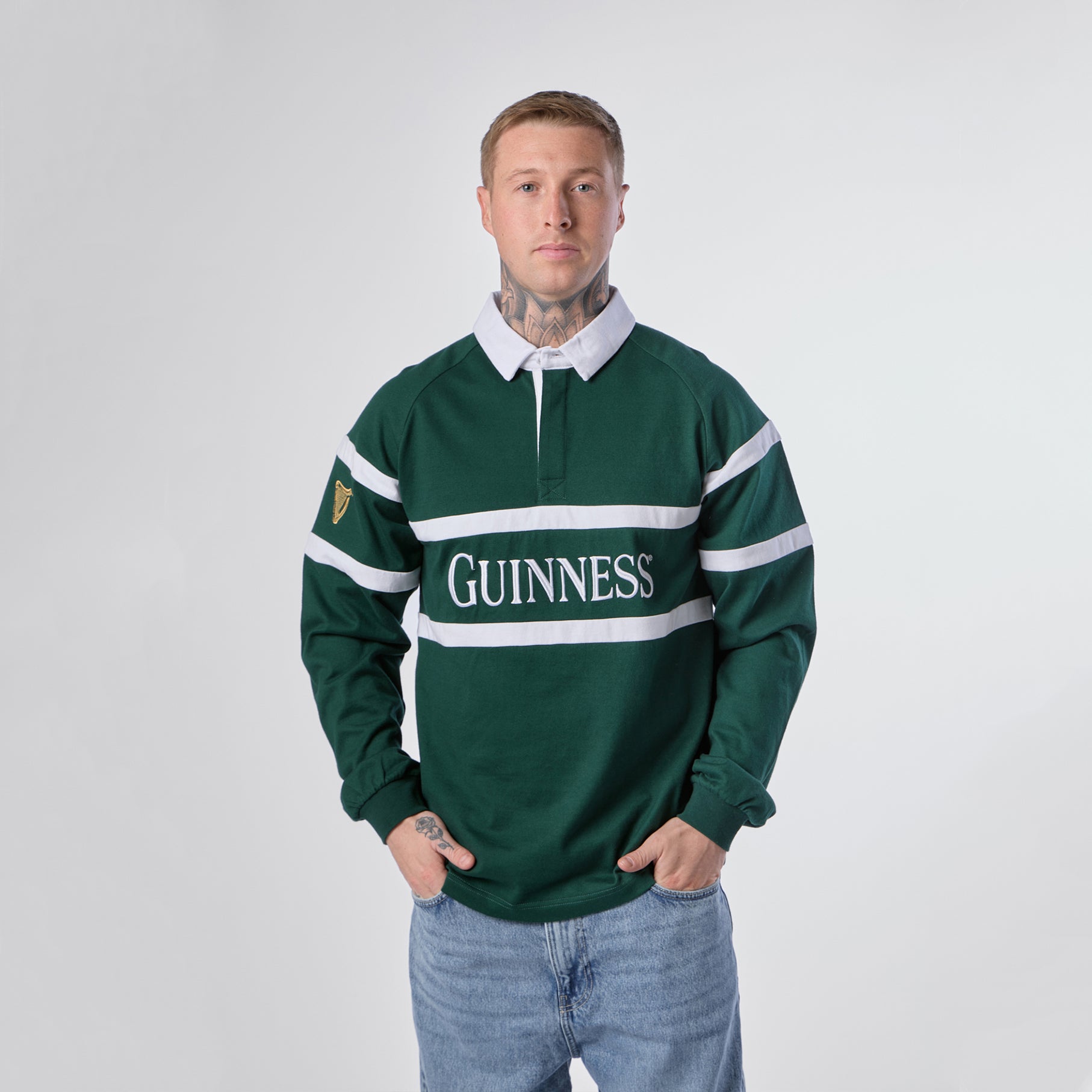 A person wearing the Guinness Green Traditional Rugby Jersey by Guinness, featuring white stripes and a white collar, stands facing forward with hands in jeans pockets against a plain background.