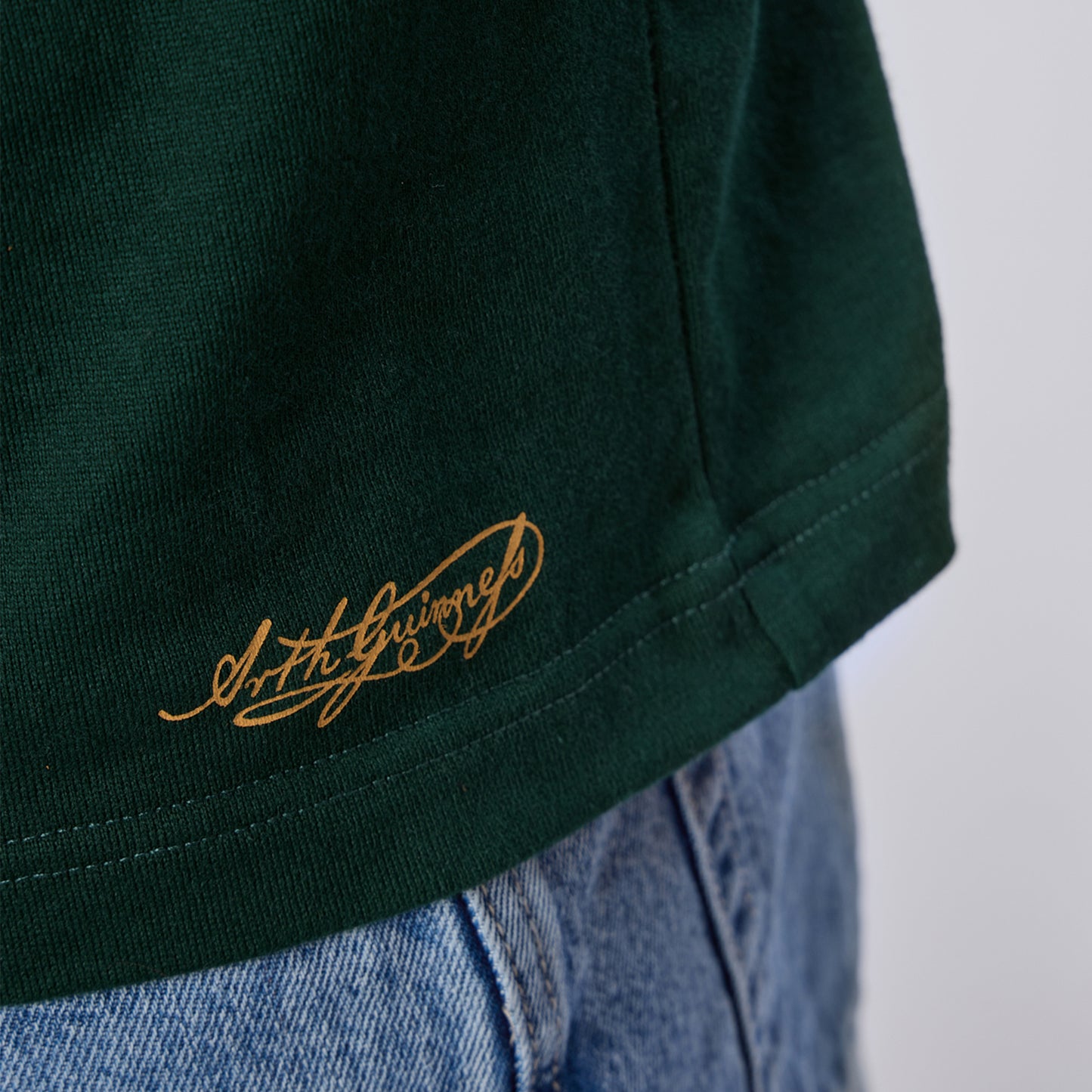 Close-up of the hem of the Guinness Green Traditional Rugby Jersey by Guinness, featuring gold script text, worn over light blue denim jeans.