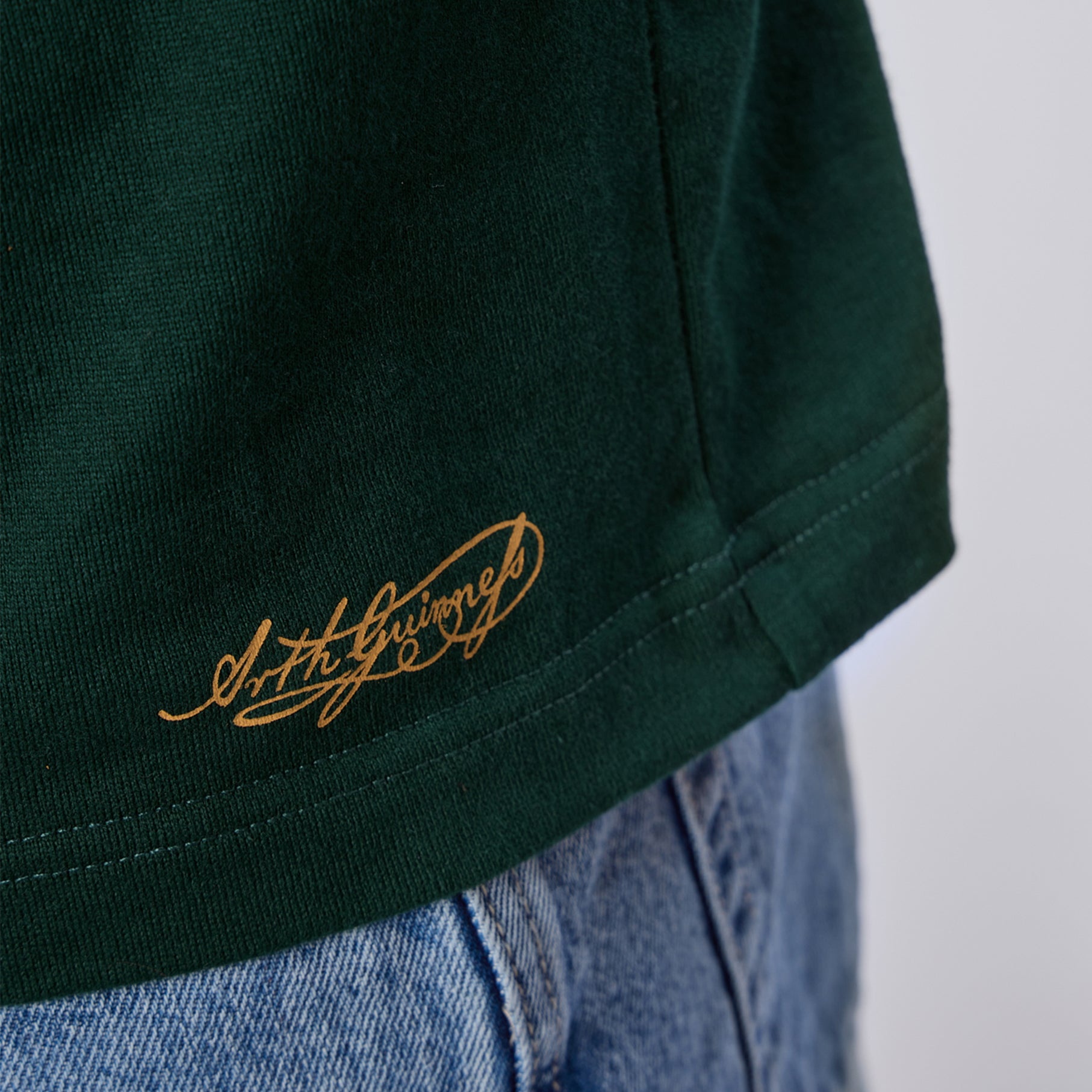 Close-up of the hem of the Guinness Green Traditional Rugby Jersey by Guinness, featuring gold script text, worn over light blue denim jeans.