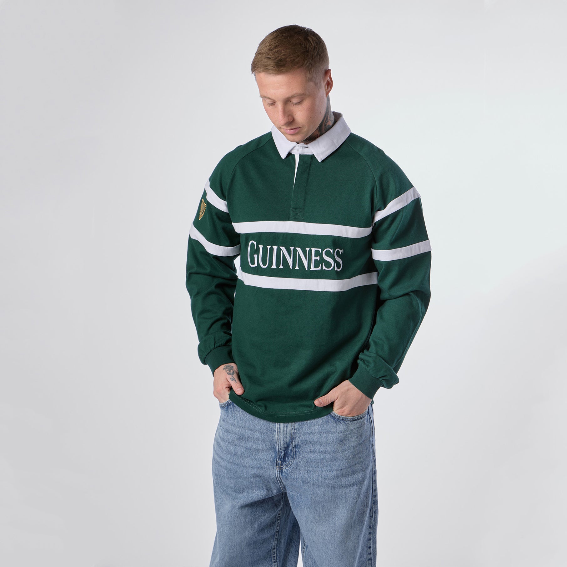 A person wearing the Guinness Green Traditional Rugby Jersey with light blue jeans stands against a plain white background, looking down.