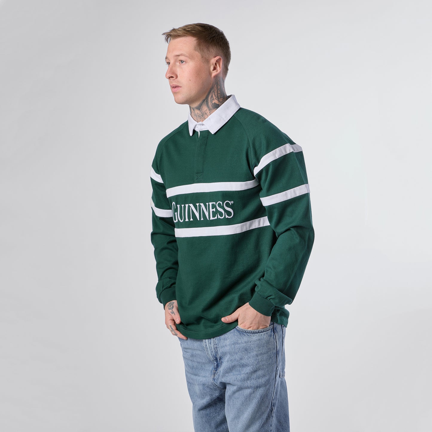 A person stands facing left, wearing the Guinness Green Traditional Rugby Jersey by Guinness, featuring “GUINNESS” text across the front, paired with light blue jeans and a white collar, set against a plain light background.