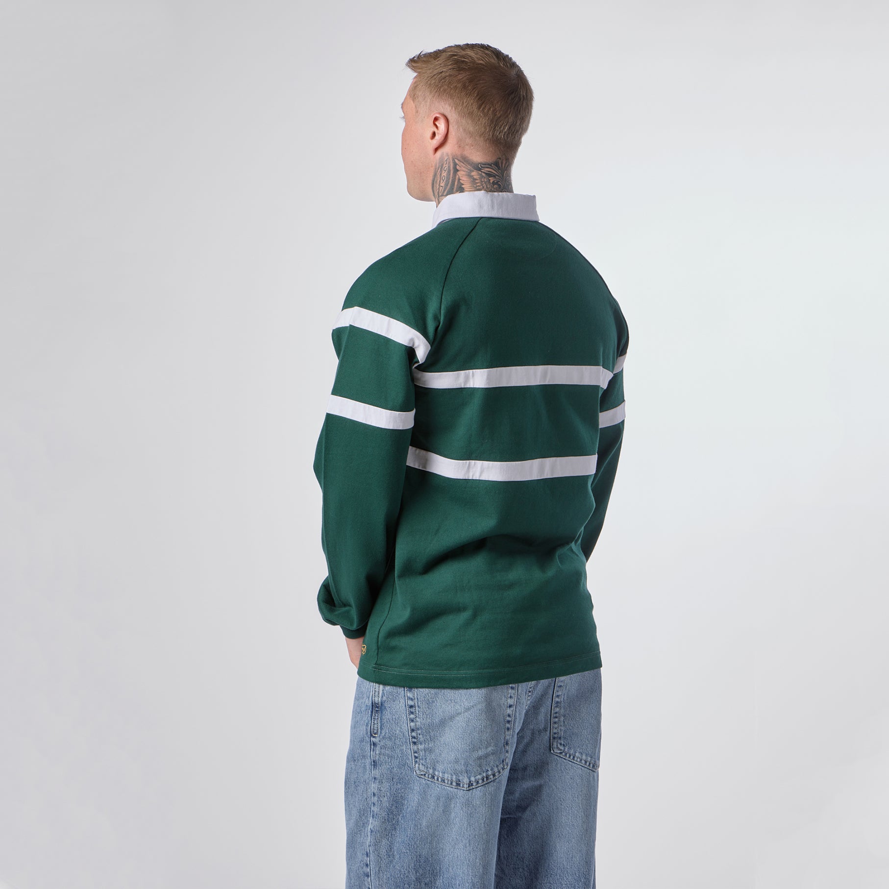 A person with short hair, wearing the Guinness Green Traditional Rugby Jersey by Guinness and blue jeans, is standing with their back to the camera against a plain background.