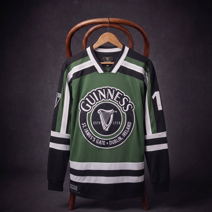 A Guinness Green & White Hockey Jersey with black accents, featuring the classic “Guinness” brand name and iconic harp logo on the front, as well as the number 17 on the sleeve, is displayed on a chair against a dark background. This bold and distinctive jersey highlights tradition for fans of both hockey and Guinness.