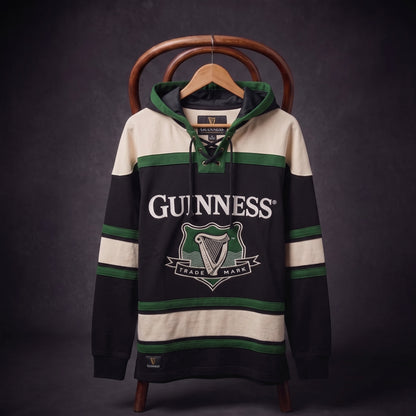 The Guinness Green Hockey Style Hooded Sweatshirt features black, green, and cream colors with a lace-up collar and the Guinness logo on the chest. Displayed on a hanger and wooden chair against a dark backdrop, this standout hoodie blends classic hockey-inspired sportswear with distinctive Guinness brand heritage.