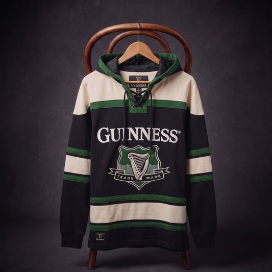 The Guinness Green Hockey Style Hooded Sweatshirt features black, green, and cream colors with a lace-up collar and the Guinness logo on the chest. Displayed on a hanger and wooden chair against a dark backdrop, this standout hoodie blends classic hockey-inspired sportswear with distinctive Guinness brand heritage.