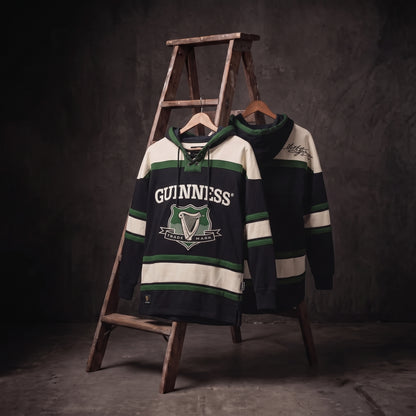 Two Guinness Green Hockey Style Hooded Sweatshirts, featuring the iconic “Guinness” name and harp logo in green, black, and cream, hang on a wooden ladder against a dark, textured background—a perfect showcase of distinctive Guinness merchandise.
