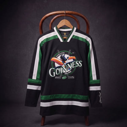 Displayed on a hanger over a wooden chair, the Guinness Toucan Hockey Jersey Black and Green features bold black and green colors with white stripes and the iconic Guinness logo showcasing the toucan. This stylish jersey from Guinness blends classic hockey design with legendary branding, perfect for fans of both the game and the brand.