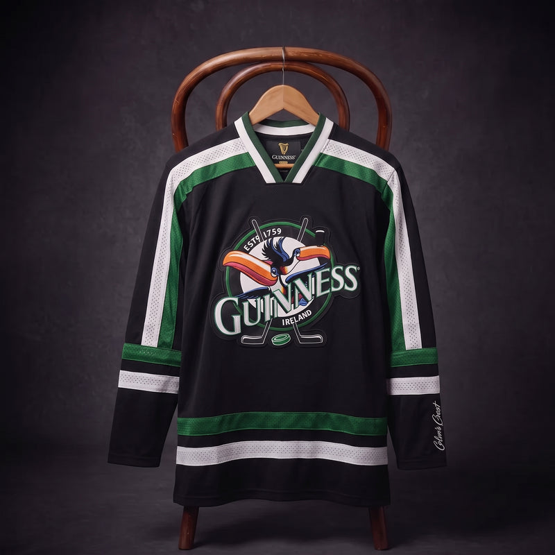 Displayed on a hanger over a wooden chair, the Guinness Toucan Hockey Jersey Black and Green features bold black and green colors with white stripes and the iconic Guinness logo showcasing the toucan. This stylish jersey from Guinness blends classic hockey design with legendary branding, perfect for fans of both the game and the brand.